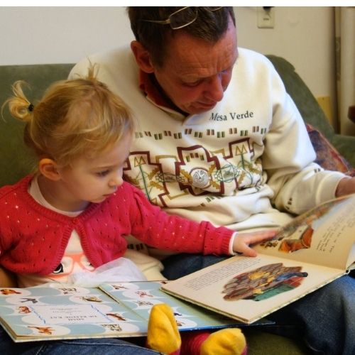 A man is reading a book to a small girl, the girl is reaching her hand out to the book.