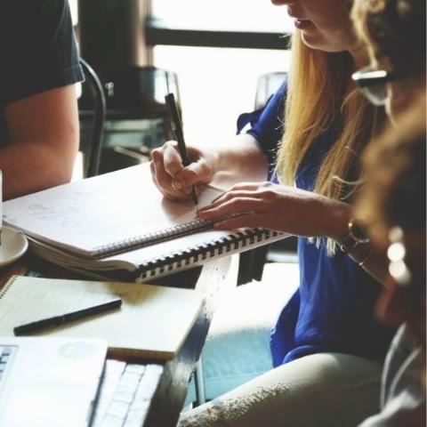 People sitting around a table, one of the women is writing in a book.